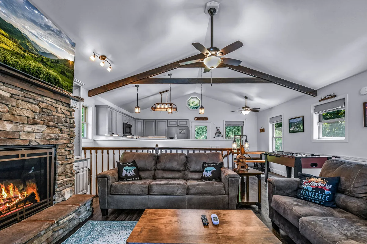 This image captures the main living area with a stone fireplace, comfortable seating, exposed wooden beams, and an open view into the kitchen area. A flat-screen TV and hints of a foosball table are visible, reinforcing the sense of warmth, entertainment, and the open-concept flow described.