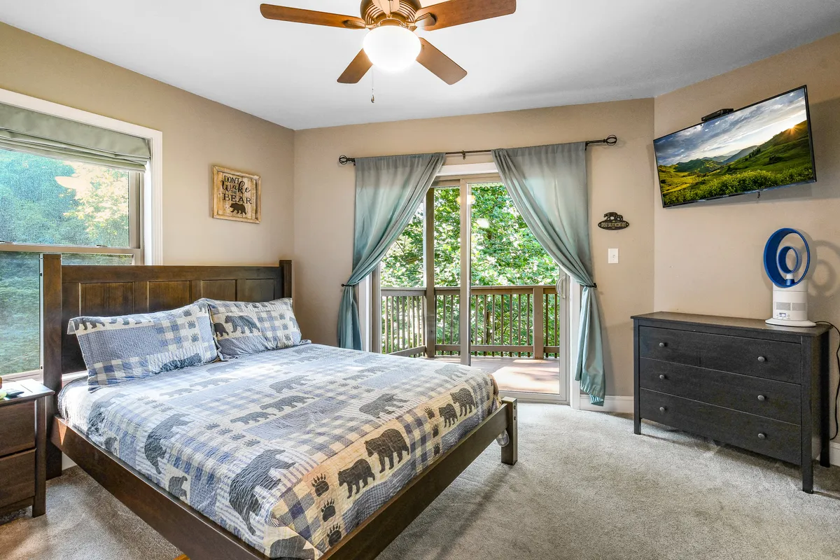 This image showcases a queen bed with bear-patterned bedding, a large window that frames views of surrounding trees, a wall-mounted television, and a dark wooden dresser. The room feels airy and comfortable, with natural light and cozy decor elements.