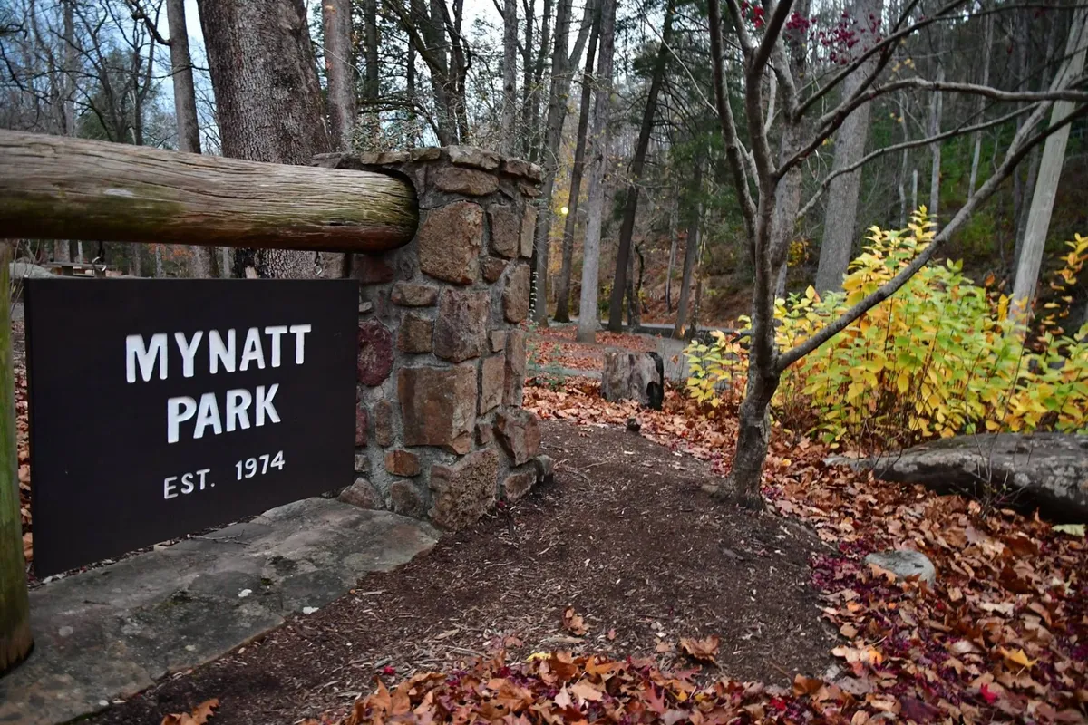The image features the welcoming sign for Mynatt Park, surrounded by colorful fall trees and a peaceful pathway perfect for walking or picnics. The park atmosphere is tranquil yet inviting, ideal for family outings or quiet reflection.