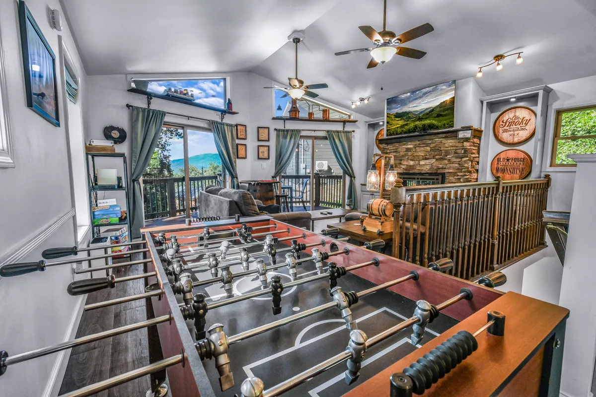 A lively game area featuring a foosball table as the focal point, flanked by comfortable seating and large windows that let in plenty of natural light. The cozy stone fireplace in the background adds warmth and ambiance, inviting guests to relax and socialize after a competitive game.