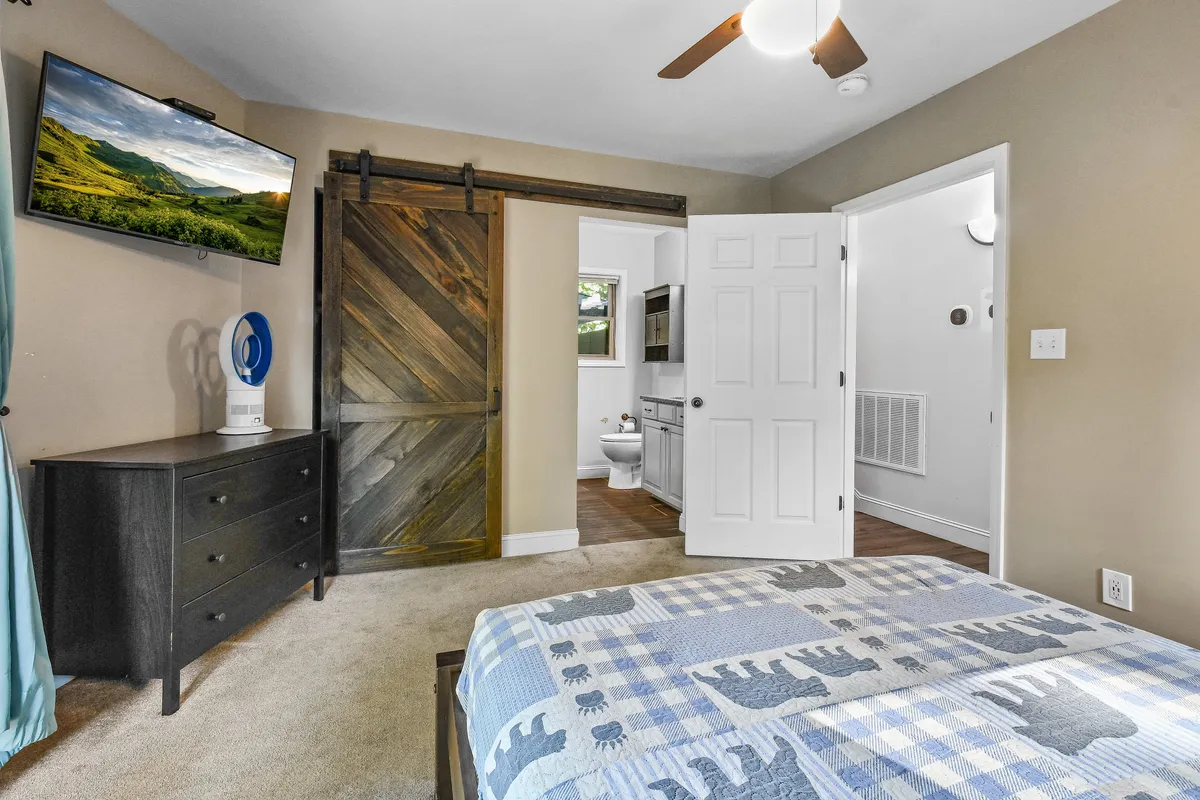 This image features a welcoming bedroom with a queen bed draped in a patterned blanket, a wooden sliding door, a wall-mounted television, and a dresser. The mood is homely and inviting, ideal for relaxation.